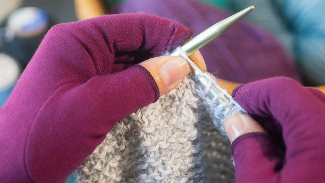 Hand of a woman with arthritis wearing Plum Purple Compression Gloves while knitting.