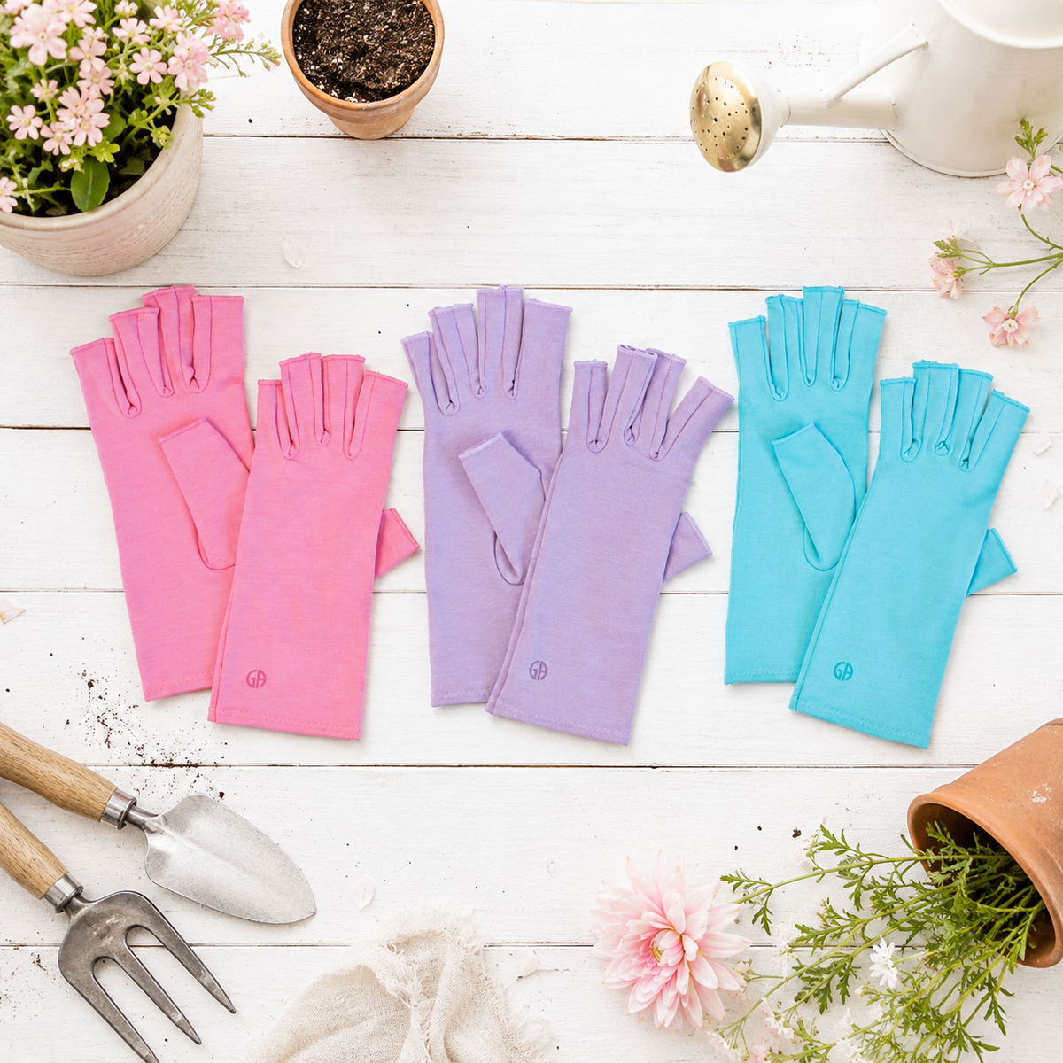 Three pairs of compression gloves in coral pink, dusky lilac, and aqua blue are arranged on a white wooden surface with gardening props including pots, flowers, a watering can, and hand tools.