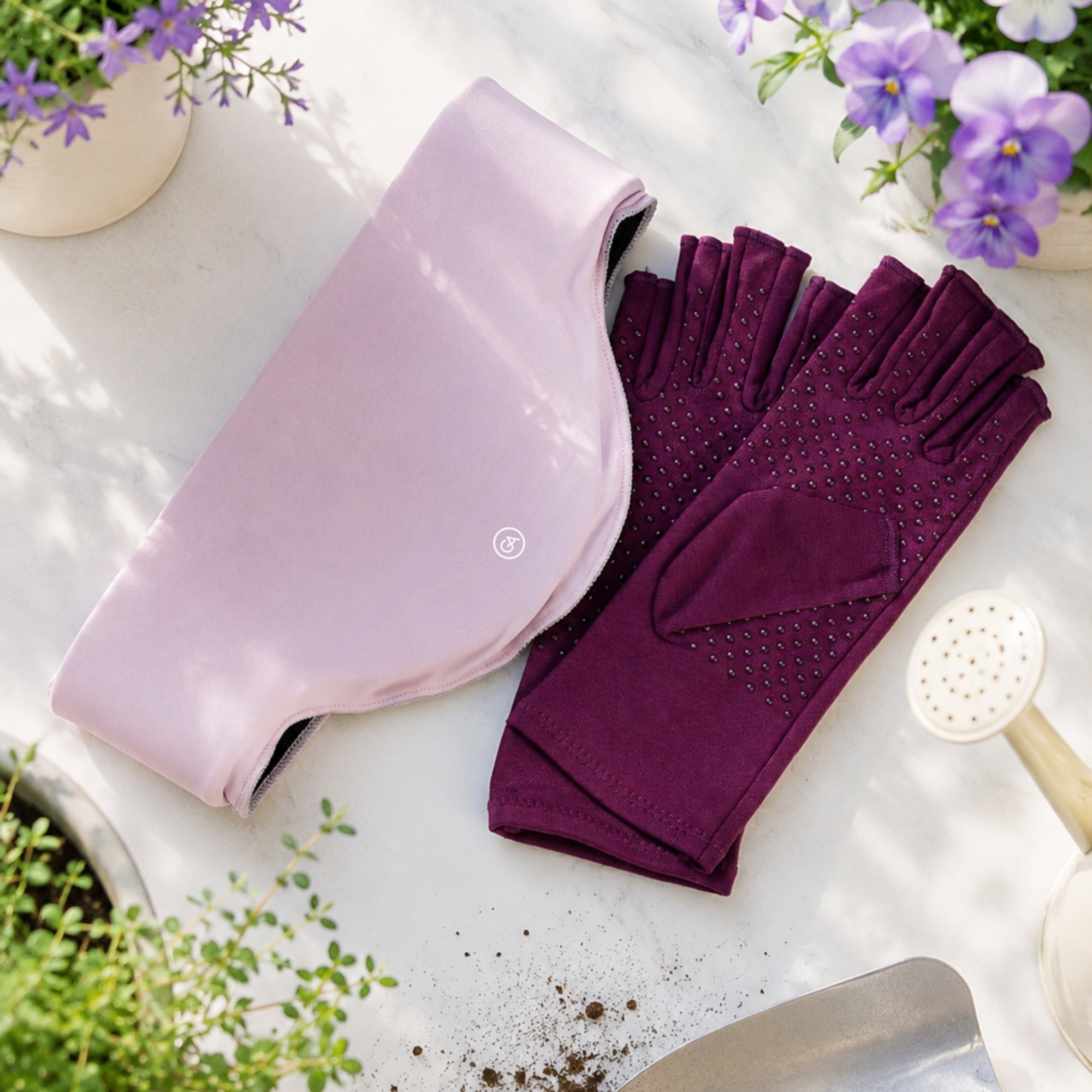 A pair of Plum Purple Extra Grip Compression Gloves with silicone dots and a matching Pastel Pink Cooling Neck Wrap are arranged on a white surface with gardening props and purple flowers around them.