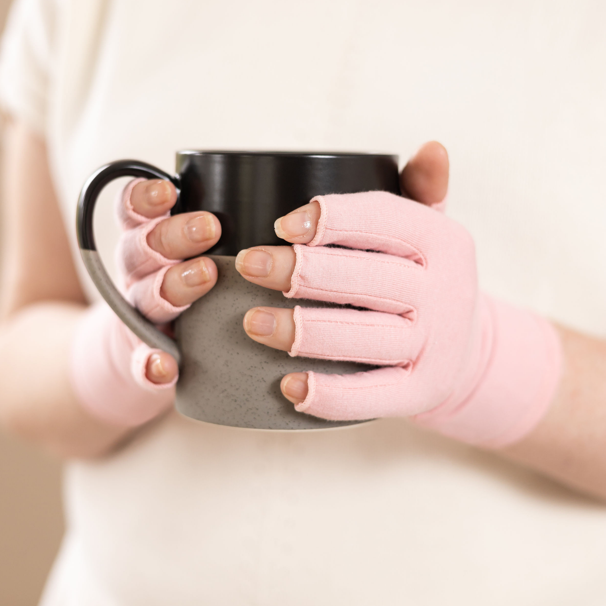 Close-up of the hands of a woman with arthritis, wearing Ballet Pink Compression Gloves, and holding a mug of turmeric tea