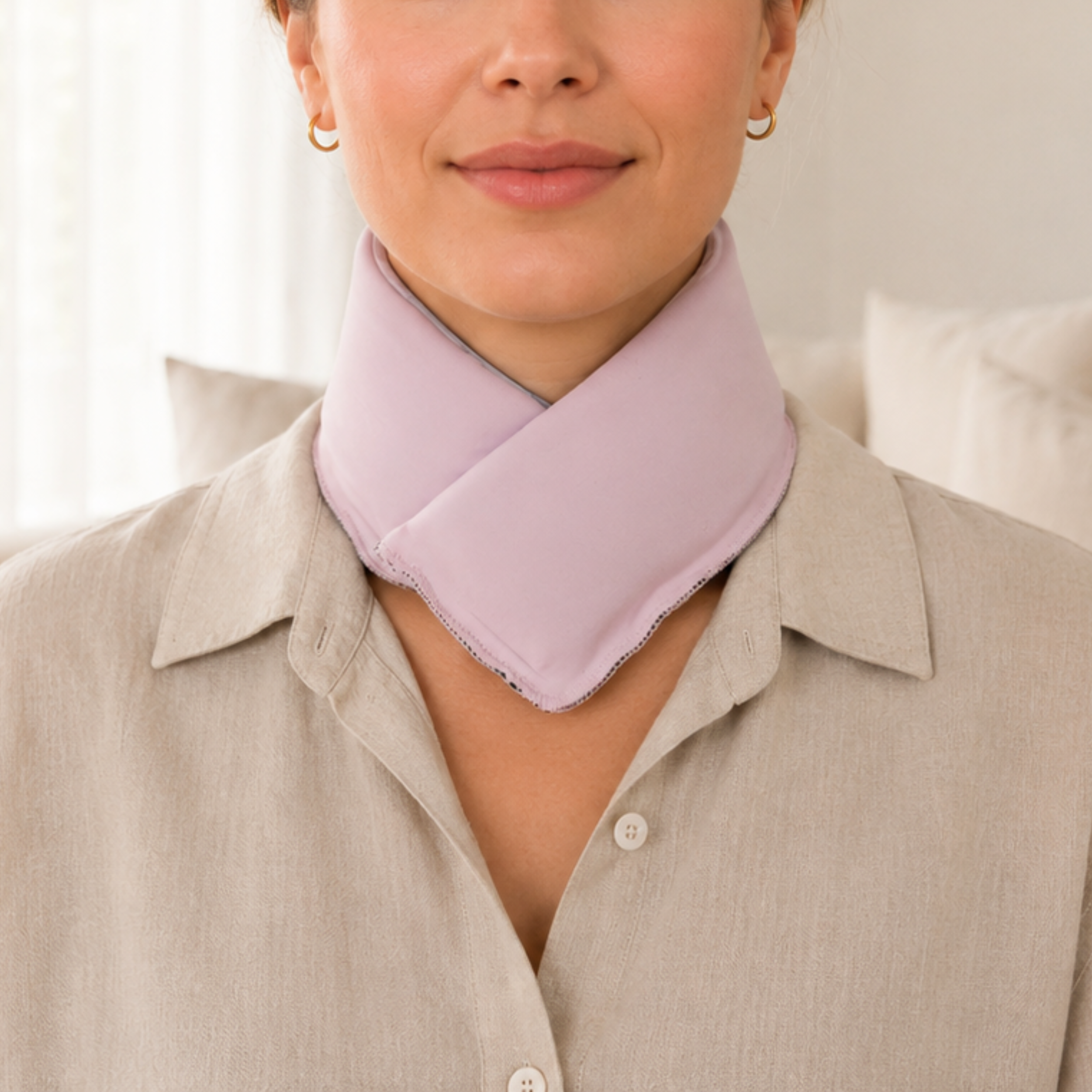 A close-up of a woman wearing a pastel pink cooling neck wrap over a light beige button-up shirt, with a softly lit indoor background.