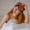 A woman with arthritis wearing a brown eyemask sitting in white blanket wakes up with insomnia painsomnia she will try wearing compression gloves to sleep / arthritis gloves in bed