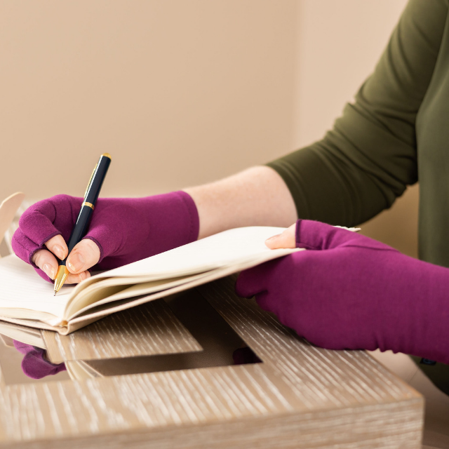 A close-up of hands of a woman with arthritis, wearing Plum Purple Compression Gloves, holding a black-golden pen and writing notes in her sketchbook.