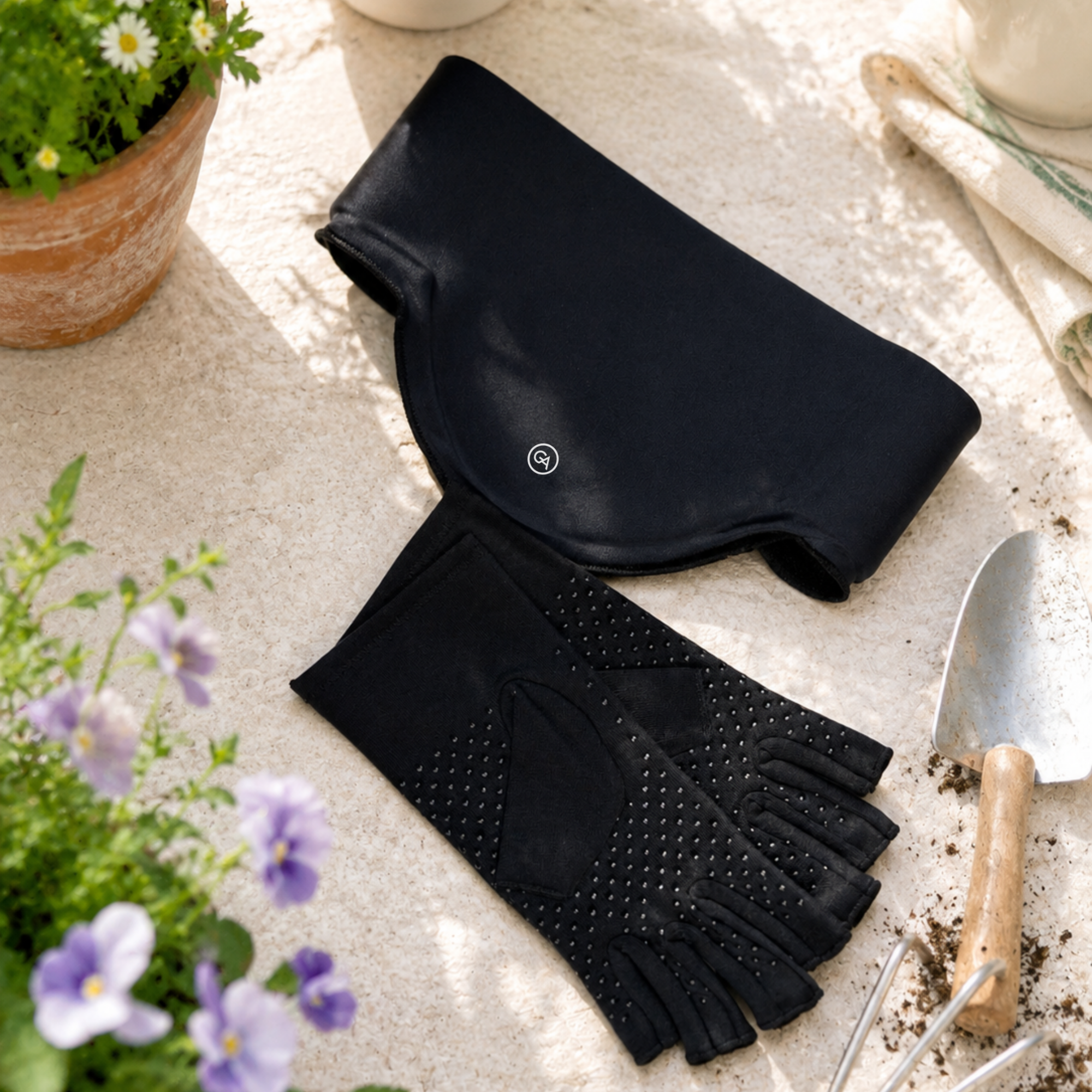 A pair of Classic Black Extra Grip Compression Gloves with silicone dots and a matching Jet Black Cooling Neck Wrap laid out on a light stone surface with gardening props including a trowel, soil, flowers, and a potted plant.