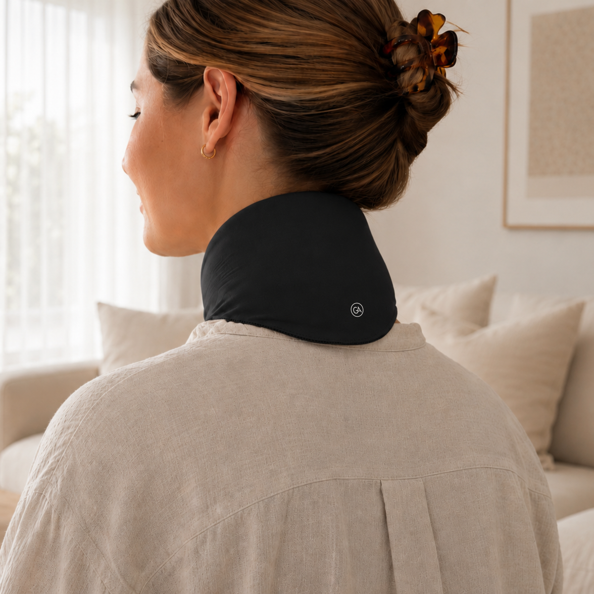 A woman shown from behind wearing a jet black cooling neck wrap in a bright, modern living room setting.