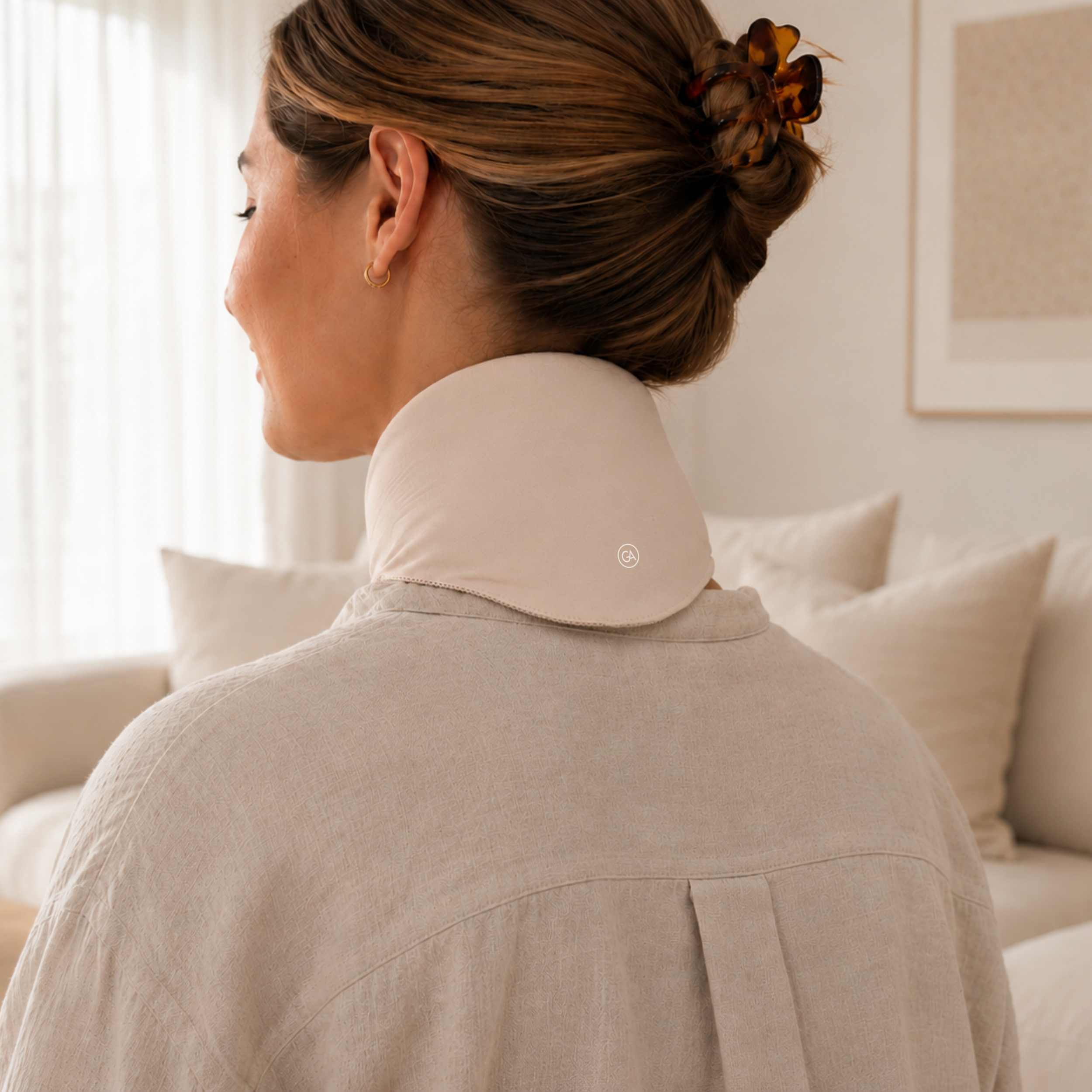 A woman shown from behind wearing a light beige linen shirt and a latte beige cooling neck wrap in a softly lit living room.