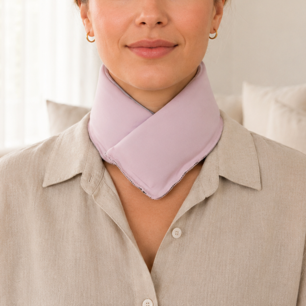 A close-up of a woman wearing a pastel pink cooling neck wrap over a light beige button-up shirt, with a softly lit indoor background.