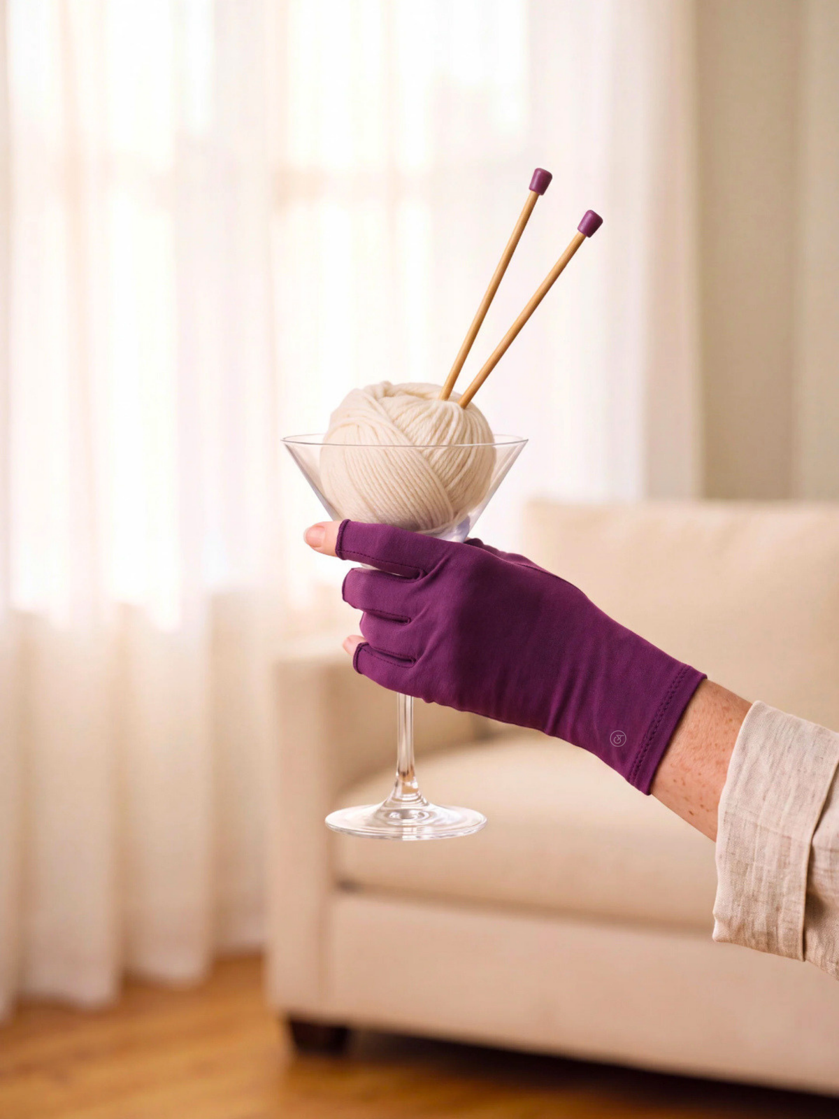 A hand wearing a purple compression gloves holding a clear glass filled with cream-colored yarn and two wooden knitting needles, set against a softly blurred living room background with a beige sofa and curtains.