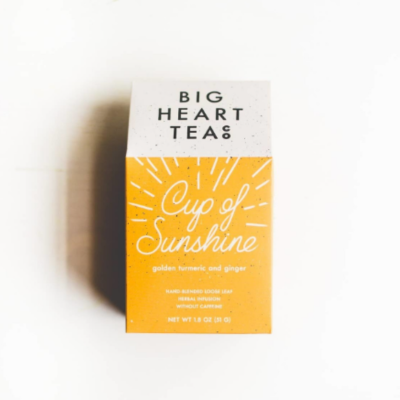 A close-up of a white and yellow tea bag box. The label says 'Big Heart Tea' and 'Cup of Sunshine'; ingredients listed: golden turmeric and ginger.